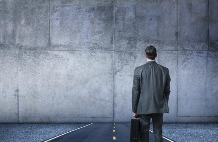 A businessman, with his back to the camera while holding a briefcase, stands in the middle of a road that runs directly into a large concrete wall.